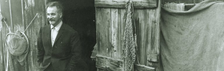 Photographie en noir et blanc de Georges Brassens devant une cabane de pêcheur en bois.