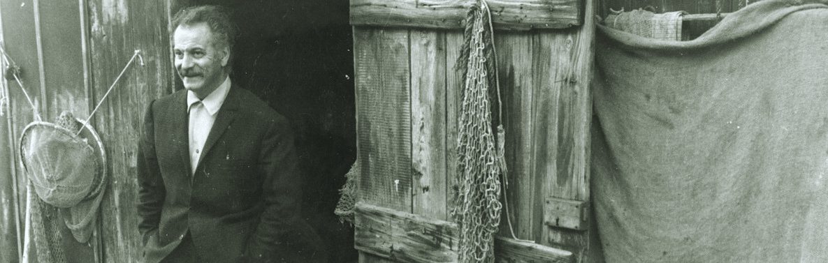Photographie en noir et blanc de Georges Brassens devant une cabane de pêcheur en bois.