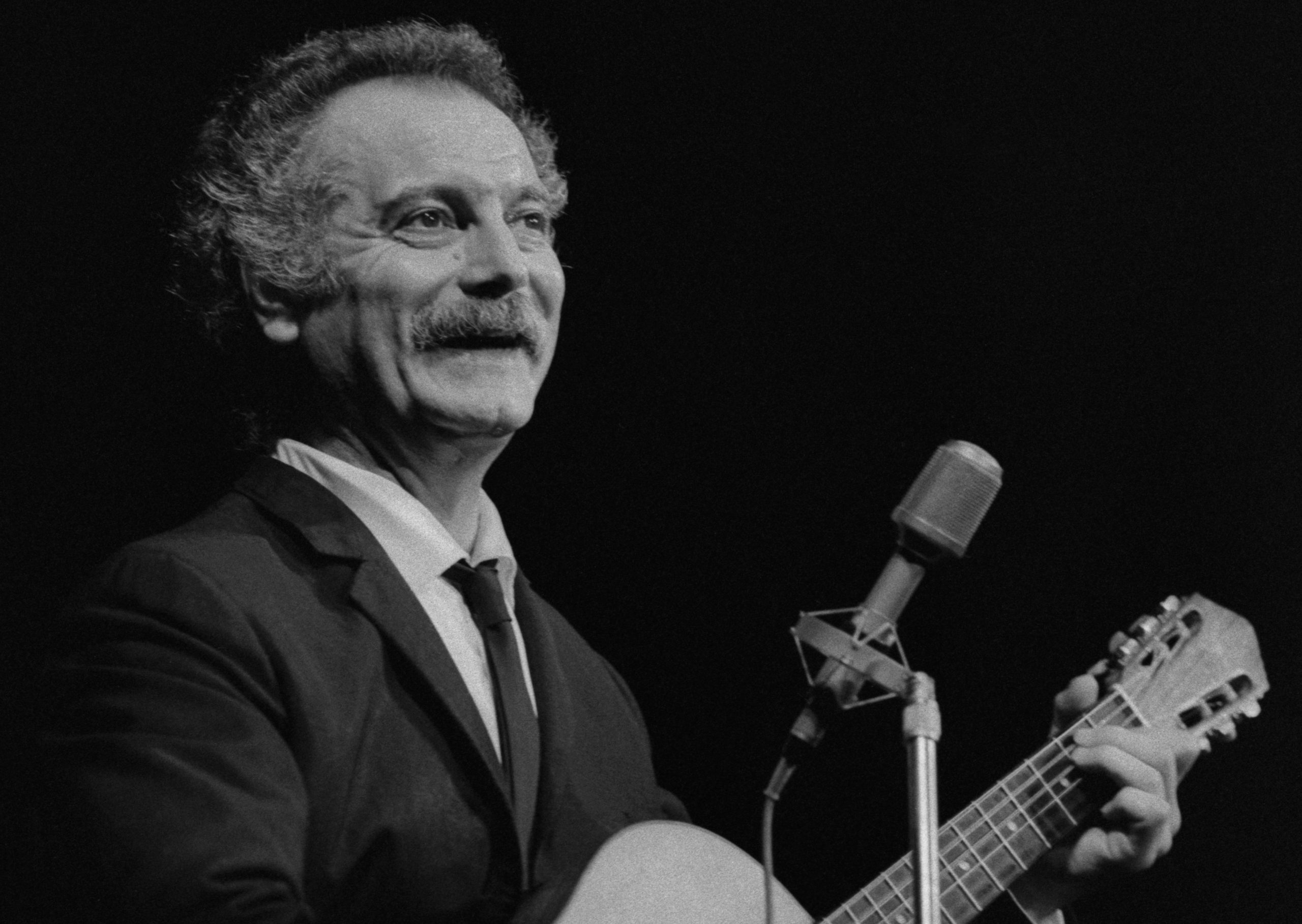 Photographie de Georges Brassens souriant en noir et blanc. En concert avec sa guitare devant un micro